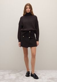 Dunkel gestreifter Rollkragenpullover kombiniert mit einem karierten Minirock mit goldenen Knöpfen und Taschen. Das Model trägt schwarze Slipper. Neutrale Hintergrund.