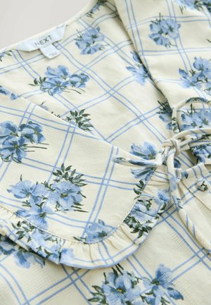 Blusa floreale con tessuto giallo decorato da motivi di fiori blu e un design a griglia, colletto arricciato e dettaglio con fiocco nella parte anteriore.