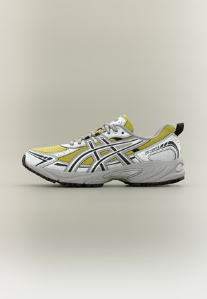 Atletisk løpesko med hvite, gule og svarte detaljer, grå såle, svart mønster og drasnor ved hælen på beige bakgrunn.