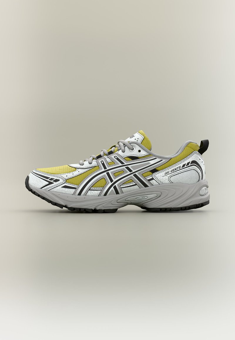 Sportlicher Laufschuh mit weißen, gelben und schwarzen Details, grauer Sohle, schwarzer Lauffläche und Zuglasche an der Ferse auf beige Hintergrund.