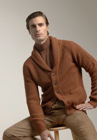 Cárdigan de punto acanalado marrón con cuello de chal, adornado con botones negros y bolsillos, usado sobre un cuello de tortuga a juego, acompañado de pantalones marrón claro.