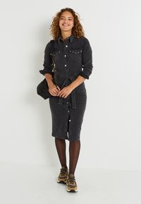 Vestido de camisa de mezclilla negra con mangas largas, bolsillos frontales y cintura ceñida con cinturón. Combinado con zapatillas marrones y medias negras transparentes.