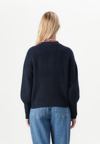 Femme aux longs cheveux blonds portant un pull en laine bleu marine et un jean bleu clair, se tenant de dos devant un fond blanc.
