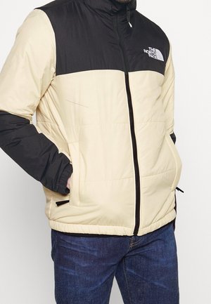 Mężczyzna w beżowo-czarnej kurtce zapinanej na zamek North Face, z rękami w kieszeniach i w ciemnoniebieskich dżinsach na jednolitym tle.