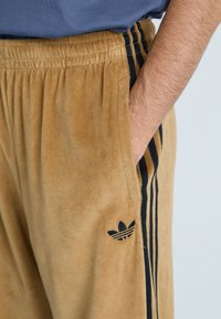 Calças de moletão de camurça bege com corte justo, cintura elástica, bolsos laterais e detalhes em preto com três riscas ao longo dos lados. Apresenta um patch com o logótipo.