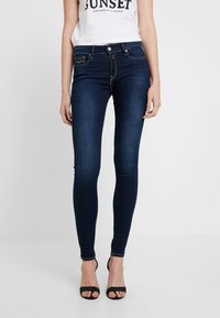 Jeans ajustados de denim azul oscuro con una textura suave, diseño de cinco bolsillos y costuras en contraste para los detalles. Llevados con tacones negros de punta abierta.