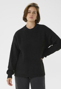 Schwarzer, gestrickter Pullover mit Rundhalsausschnitt, fallenden Schultern und langen Ärmeln; weicher Stoff und lässige Passform. Kombiniert mit dunklen Jeans.