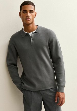 Young man wearing a gray long-sleeve polo shirt over a white undershirt, and checkered pants, standing against a beige wall.