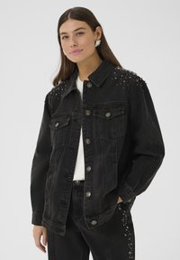 Tânără îmbrăcată într-o jachetă de denim neagră și pantaloni cu ținte argintii, cămașă albă și cercei argintii, pozând cu mâna în buzunar.