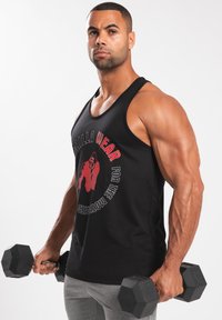 Zwarte tanktop met rode grafische tekst en een gorillatekening. Model houdt hexagonale dumbbells vast. Grijze sportbroek, gespierde armen zichtbaar.