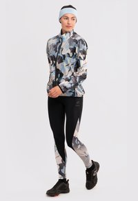 Chaqueta ligera con patrón de camuflaje en tonos de azul, gris y negro, combinada con leggings negros que tienen un diseño similar y zapatillas deportivas.