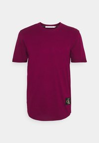 Camiseta corta de manga en burdeos con cuello redondo y un pequeño parche con el logo de Calvin Klein cerca del dobladillo inferior sobre un fondo blanco.
