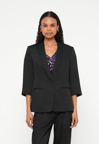 Blazer negro a medida con cuello chal, cierre de un solo botón y bolsillos laterales. Se lleva sobre una blusa morada con lentejuelas. Textura suave.