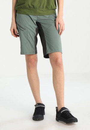 Osoba stojící na světle jednobarevném pozadí, oblečená v zelených a šedých krátkých kalhotách po kolena s černými detaily a černých sportovních botách.