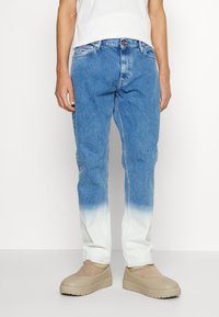 Osoba oblečená v modro-bílých ombre džínech v kombinaci s béžovými slip-on botami a bílou košilí, stojící na bílém pozadí.