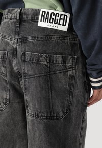 Schwarze Jeans aus Denim mit einer verblassten Textur, ausgestattet mit einem weißen Etikett mit dem Logo "RAGGED JEANS", sichtbaren Gesäßtaschen und einer hohen Taille.