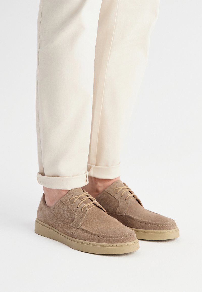 Brązowe zamszowe buty z okrągłym czubkiem, niskim profilem i beżowymi gumowymi podeszwami. Posiadają jasnobrązowe sznurowadła oraz przeszycia na cholewce.