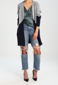 Gestrickter Cardigan in Grau und Marineblau, kombiniert mit einem grünen Satintop. Dazu werden blaue, distressed Jeans mit ausgefransten Säumen und hochhackige Schuhe getragen.