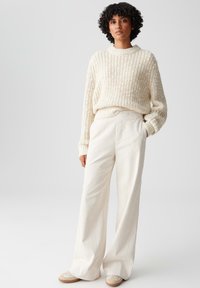 Maglione spesso crema con una texture a coste, abbinato a pantaloni a gamba larga crema. Le sneakers presentano tonalità neutre con leggeri accenti.