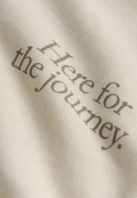 Primo piano di un tessuto beige con le parole "Here for the journey." stampate in carattere serif.