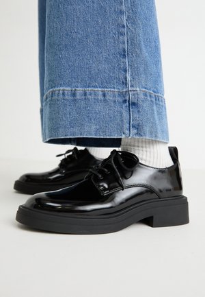 Sorte skinnende snørestøvler med plateau, båret med hvide ribstrikkede sokker og brede blå denimbukser, der er forkortet over anklen.