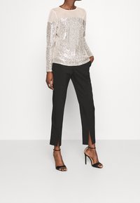 Pailletten-Oberteil mit einem durchsichtigen beigen oberen Bereich; kombiniert mit schwarzen Hosen mit Schlitzen und schwarzen Knöchelriemen-Absätzen.