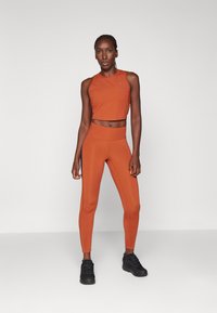 Narančasti crop top s volanima u kombinaciji s narančastim tajicama visokog struka. Uparen s crnim sportskim cipelama. Glatka, rastezljiva tkanina.