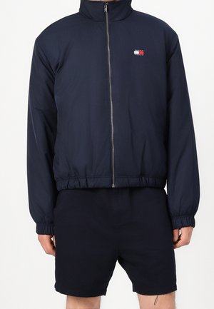 Mężczyzna noszący granatową kurtkę na zamek z małym logo Tommy Hilfiger na piersi oraz dopasowane granatowe szorty, ręce swobodnie opuszczone wzdłuż ciała.