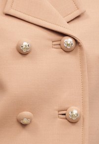 Μπεζ διπλόκουμπο σακάκι με τέσσερα κουμπιά καλυμμένα με ύφασμα που φέρουν λευκά και χρυσά λουλουδένια σχέδια.