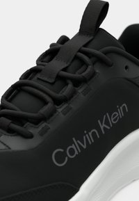 Μαύρο αθλητικό παπούτσι με λεία υφή, στρογγυλή μύτη, υφασμένες κορδόνια και λευκή σόλα. Φέρει το λογότυπο "Calvin Klein" σε γκρι.