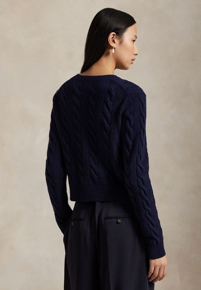 Kvinde med langt mørkt hår iført navyblå kabelstrikket sweater og mørke vidde bukser, stående sidelæns mod en ensfarvet neutral baggrund.