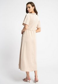 Długa, beżowa sukienka z krótkimi rękawami, teksturowany materiał z małymi kropkami, taliowana w pasie i rozkloszowany dół. Noszona z różowymi szpilkami.