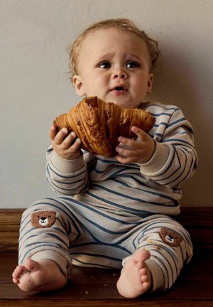 Toddler in striped pajamas with bear patches sitting on floor, holding a large croissant with crumbs around.