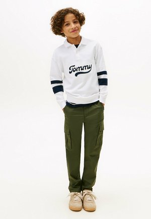 Witte poloshirt met lange mouwen en marineblauwe strepen, voorzien van het "Tommy"-logo, gecombineerd met olijfgroene cargobroeken en beige sneakers.