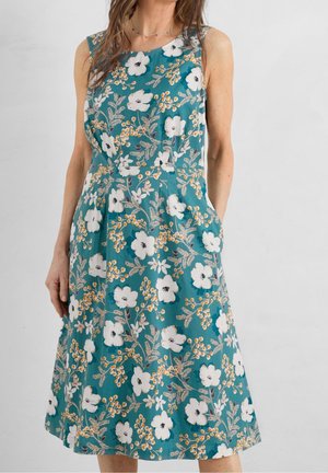 Robe sans manches couleur sarcelle avec un motif floral blanc et jaune portée par une personne posant devant un fond uni.