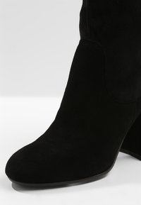 Bottine en daim noir avec bout arrondi et talon bloc sur un fond clair uni.