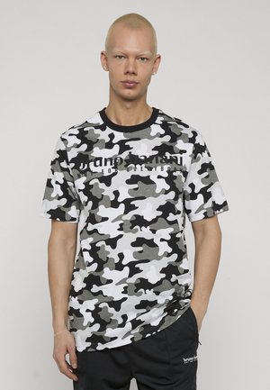 Giovane con capelli biondi corti che indossa una maglietta mimetica nera, bianca e grigia e pantaloni neri con la mano in tasca, in piedi su uno sfondo semplice.