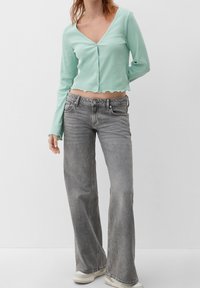 Měkký zelený zkrácený top s dlouhým rukávem, výstřihem do V a krajkovým lemem, doplněný šedými rozšířenými džíny. Bílá obuv dokončuje vzhled.