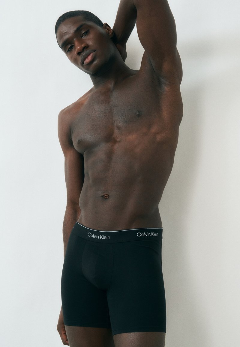 Άνδρας χωρίς πουκάμισο που φοράει μαύρα εσώρουχα Calvin Klein, στέκεται μπροστά σε έναν απλό ανοιχτό τοίχο με το ένα χέρι σηκωμένο πίσω από το κεφάλι του.