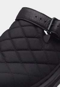 Μαύρο παπούτσι από υφασμάτινο καπιτονέ με υφή, ρυθμιζόμενο λουρί από δέρμα με ανάγλυφη επιφάνεια και μαύρη μεταλλική πόρπη.