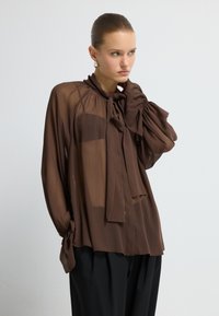 Frau mit zurückgegeltem Haar, die eine durchsichtige braune Bluse mit Halsband und schwarze Hose trägt, posiert mit einer Hand nahe ihrem Hals.