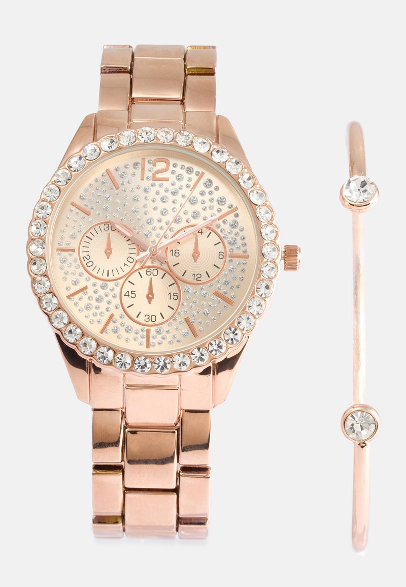 Roza zlata metalna ura z bleščečimi rhinestones na okvirju in številčnici, ki vsebuje tri podštevce. Vključuje usklajen bangle zapestnico.
