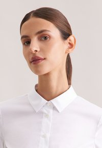 Kobieta o brązowych włosach zebranych do tyłu, ubrana w białą koszulę na guziki, patrząca lekko na bok na tle jasnego, jednolitego tła.