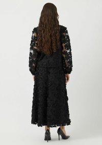 Svart kjole med blomsterbroderier, gjennomsiktige lange ermer, et luftig skjørt og frynser nederst, kombinert med sorte høyhælte sko.