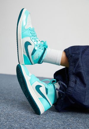 Personne portant des baskets montantes Nike turquoise et blanches, des chaussettes blanches et un pantalon bleu foncé à cordon, allongée sur un sol recouvert de moquette.
