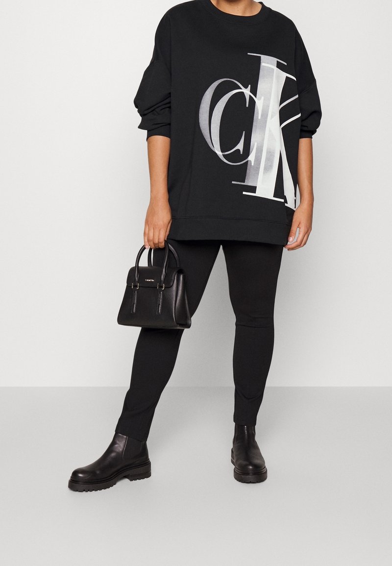 Femme portant un sweat-shirt noir oversize avec un grand logo blanc "CK", des leggings noirs, des bottes noires, tenant un petit sac à main noir.