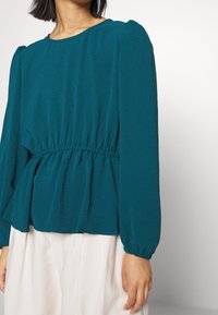Blusa teal a maniche lunghe con vita elastica. Tessuto morbido con una leggera texture. Collo rotondo e dettagli arricciati sulle spalle.