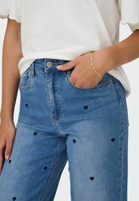 Zbliżenie osoby w jasnoniebieskich dżinsach z małymi ciemnymi sercami i białej bluzce, ręka oparta o przednią kieszeń, na której znajduje się złota bransoletka.