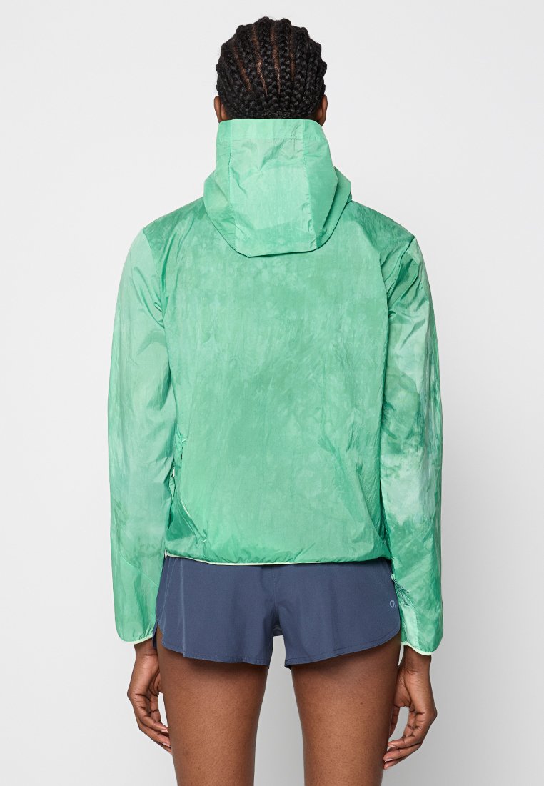 Zielona, lekka kurtka z kapturem, o gładkiej teksturze i lekko błyszczącym wykończeniu. Opadający dół i bezszwowe rękawy. Widoczne szorty poniżej.