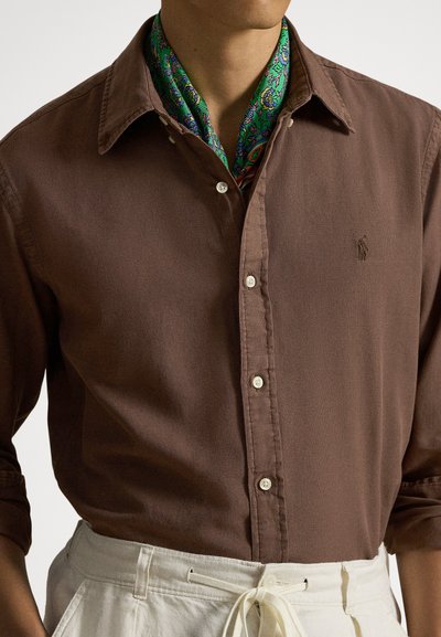 Καφέ πουκάμισο με κουμπιά από υφασμάτινη ανάγλυφη ύλη, με κλασικό γιακά και μικρό λογότυπο. Μια πράσινη φουλάρι με σχέδια είναι κρυμμένη στο γιακά.